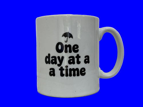 A white ceramic coffee mug with black text reading 'One day at a a time' and a small umbrella graphic above it, shown on a plain white background.